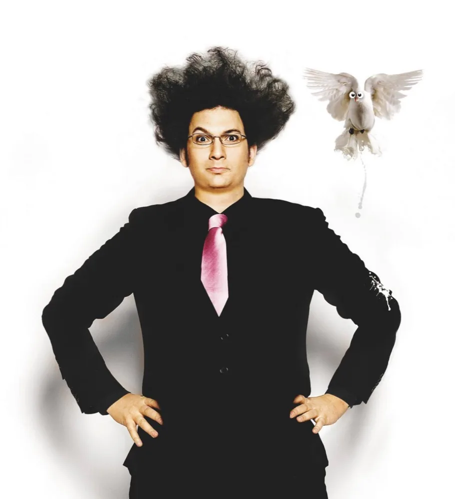 Éric Antoine en costume noir et cravate rose, cheveux hirsutes et lunettes rondes, une colombe en vol à côté de lui