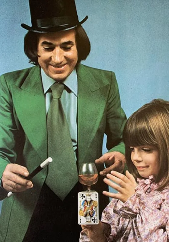 Garcimore en costume vert et chapeau haut de forme, baguette à la main, présentant une carte en équilibre sur un verre à une petite fille