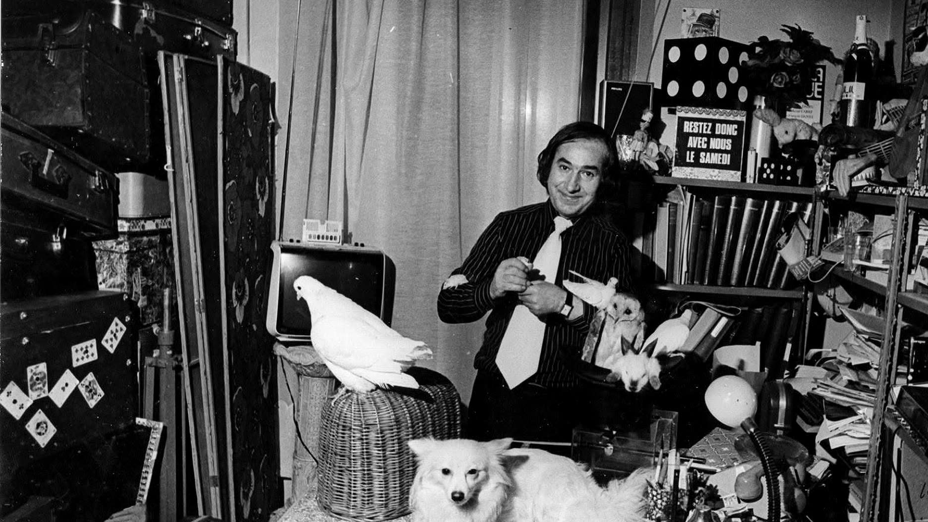 Garcimore dans son atelier, entouré de malles, d'accessoires et de livres, une colombe sur un panier, sa chienne Dolly couchée devant lui et au mur le panneau « Restez donc avec nous le samedi »