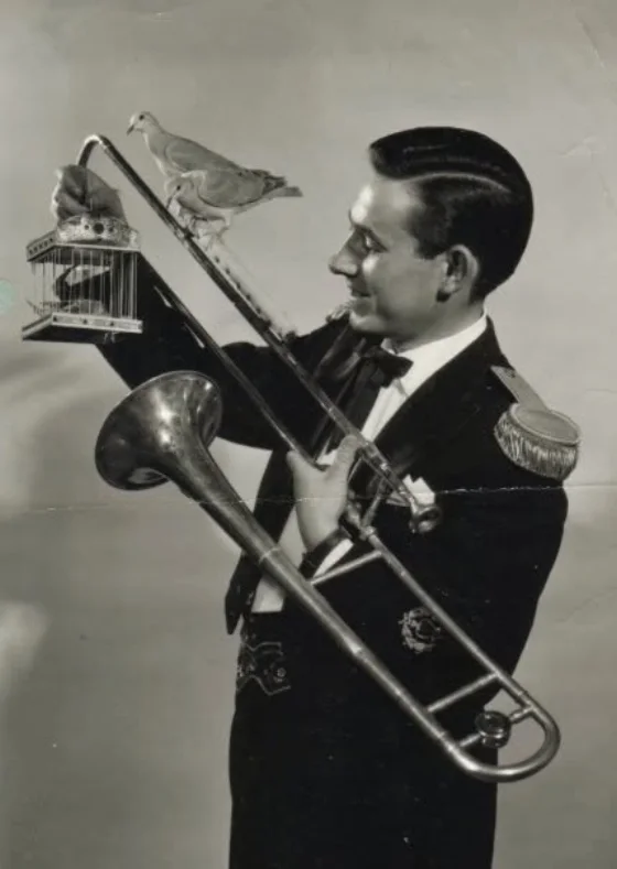 Garcimore jeune homme en veste à galons, un trombone à la main, une colombe posée sur la coulisse et une cage à oiseaux dans l'autre main