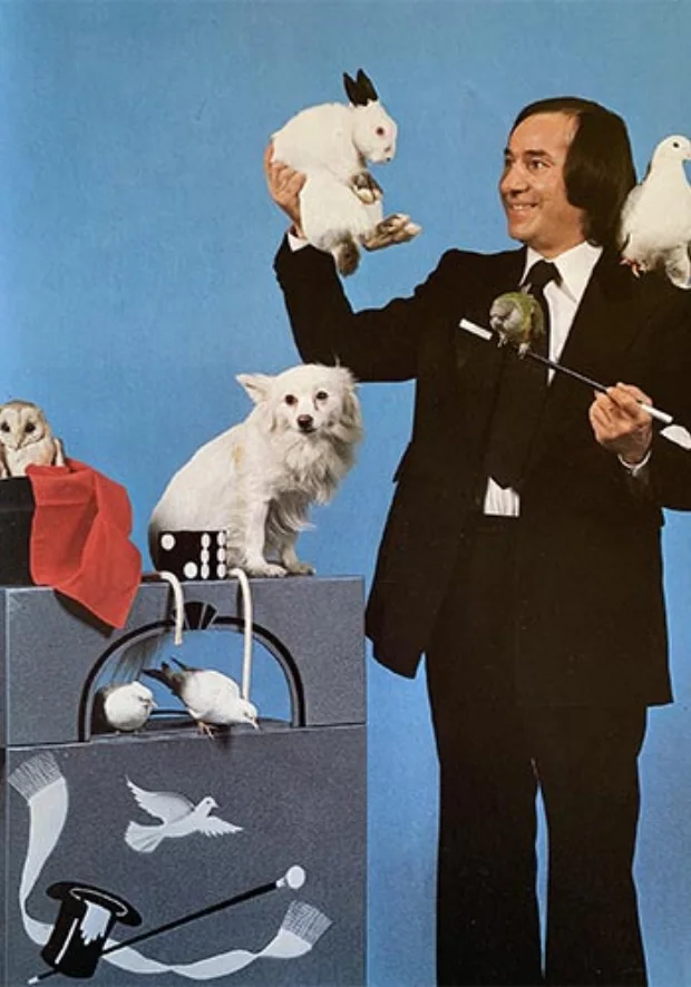 Garcimore en habit noir, un lapin blanc brandi à bout de bras, une colombe sur l'épaule, sa chienne Dolly et une chouette posées sur ses caisses de scène