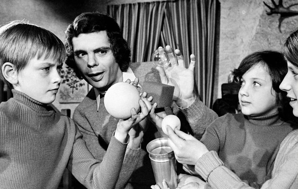 Gérard Majax jeune, en pull, présente un tour de balles en mousse et de gobelet à quatre enfants qui l'observent de très près