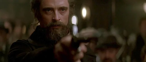 Hugh Jackman en Angier, grimé avec une barbe, braque un pistolet vers la scène depuis le public