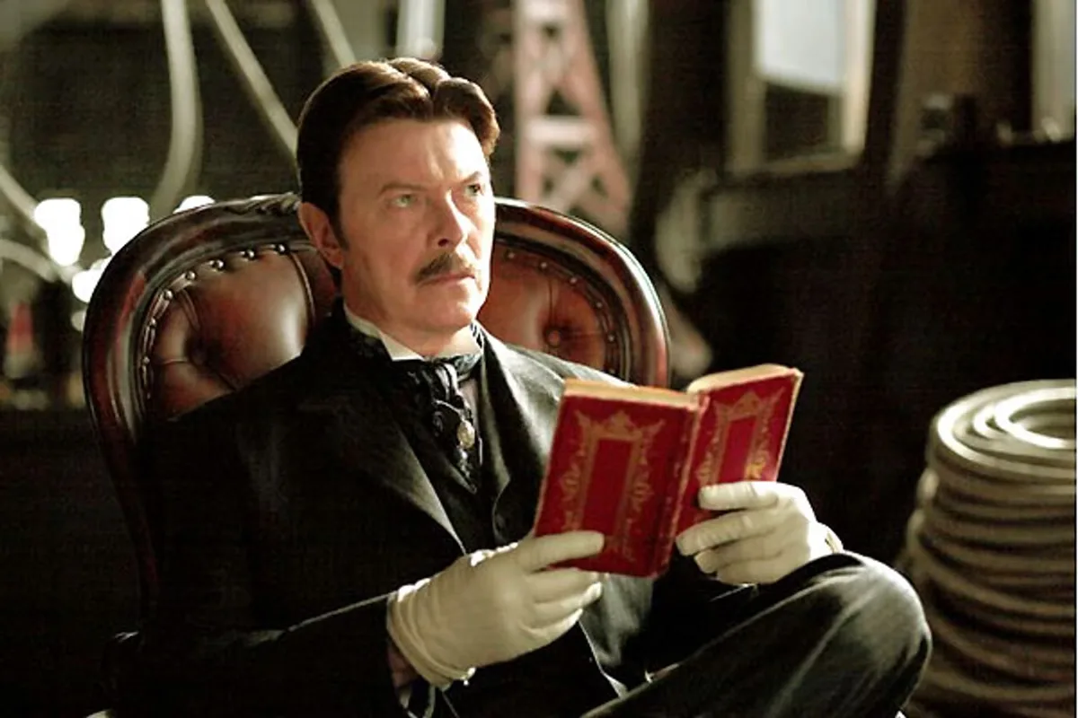 David Bowie en Nikola Tesla, assis dans un fauteuil de cuir, lisant un livre rouge, gants blancs aux mains